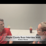 Two people seated at a table in Palm Coast, engaged in an interview The man on the left wears a red shirt, while a woman with glasses sits on the right Two drinks with straws are on the table, and a text overlay reads "Flagler County Buzz Interview With Andrew Werner